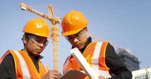 Two construction workers wearing orange safety vests and helmets, reviewing a clipboard on a construction site with cranes in the background