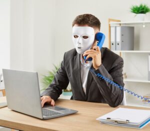 Man wearing a mask, using a phone and laptop at a desk.