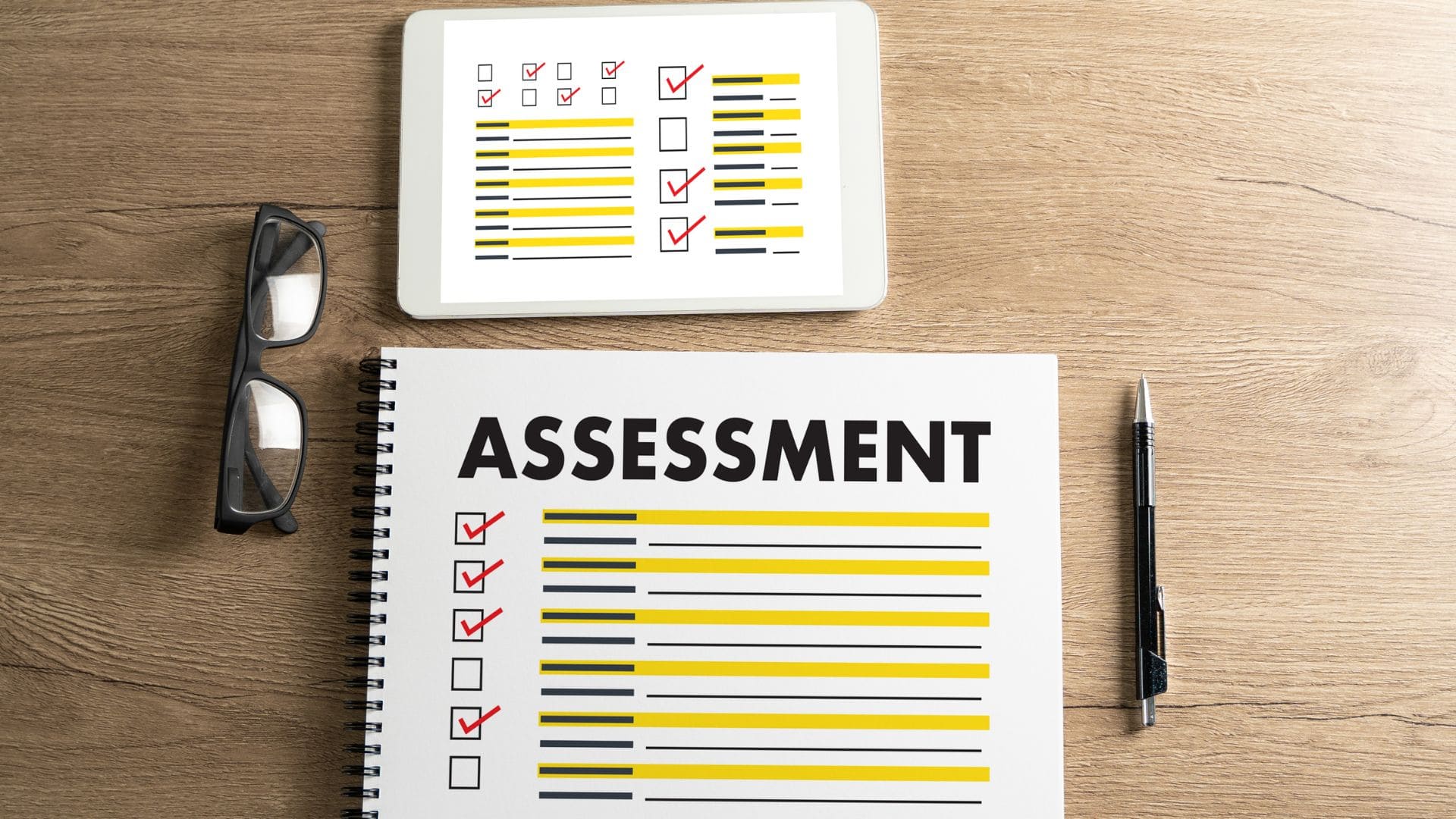 Assessment form with glasses, pen, and tablet on a wooden desk