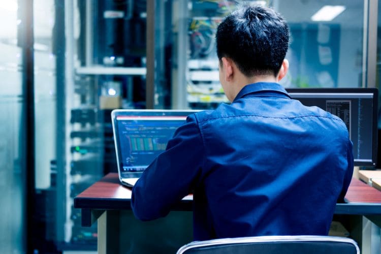 A man working on a laptop in a server room, with coding displayed on the screen.