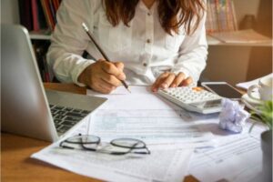 Person calculating finances with a calculator, papers, and a laptop on a desk.